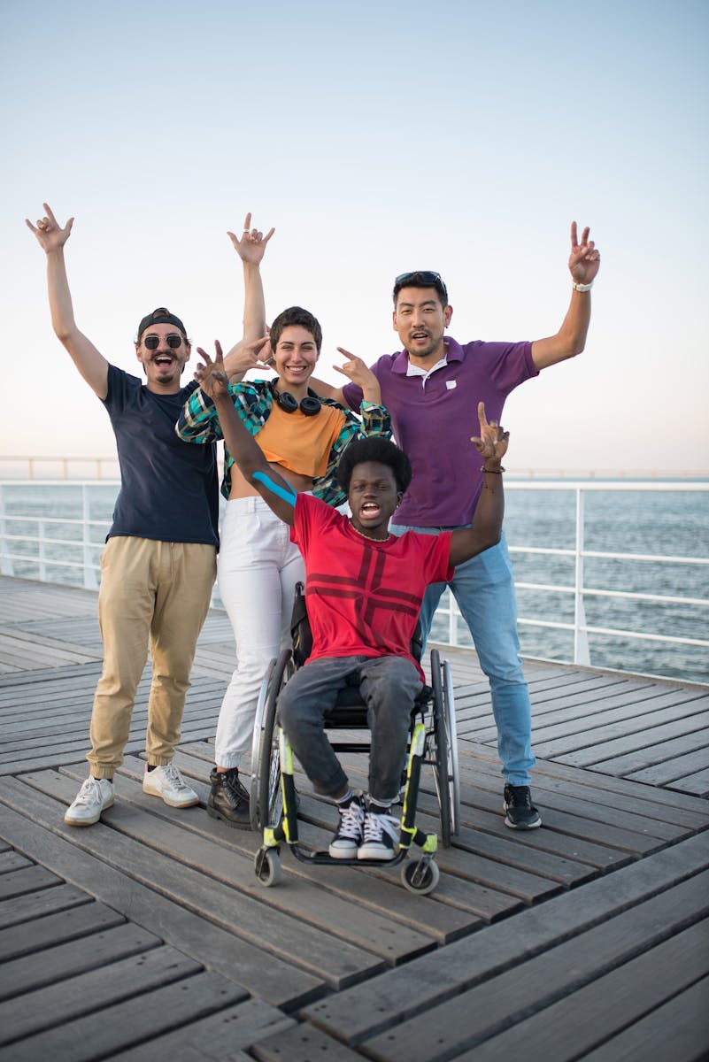 Group of diverse friends, including a person in a wheelchair, joyfully posing outdoors by the waterfront.